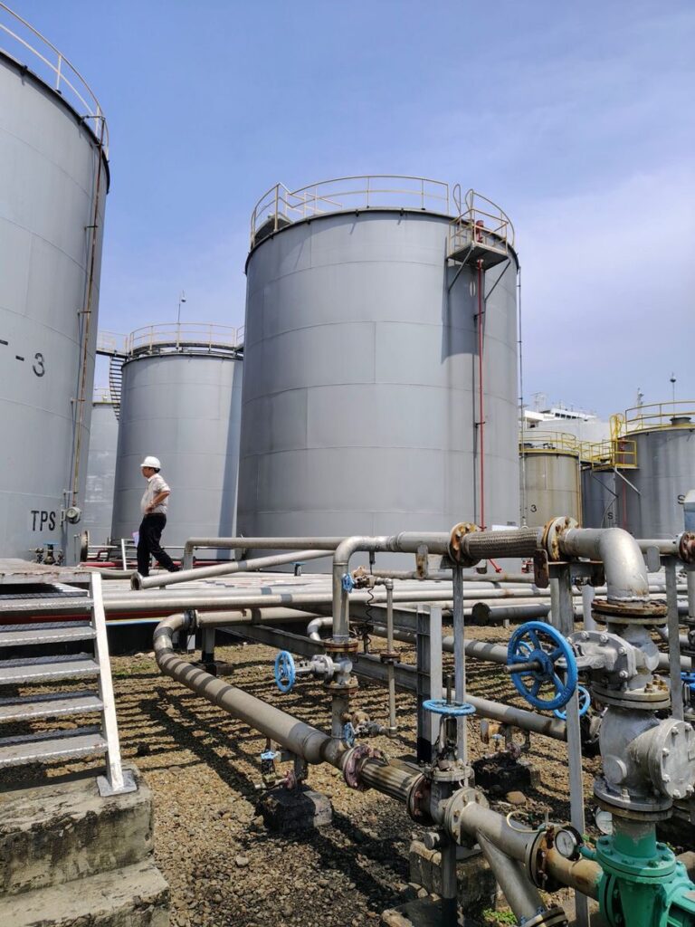 Person walking near large industrial oil storage tanks and pipelines under a clear blue sky.