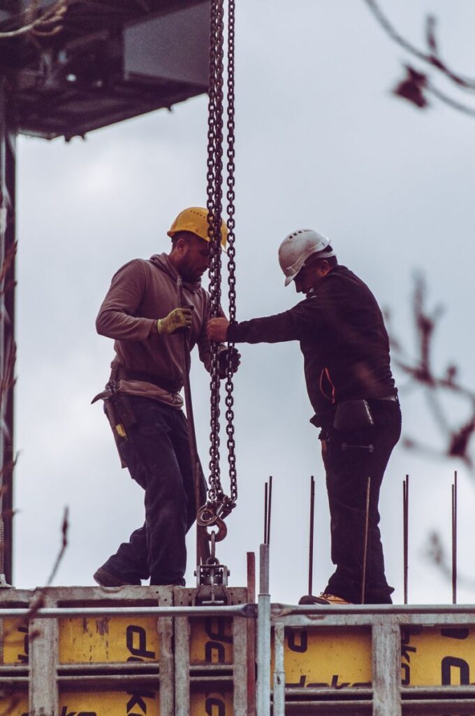 Two workers in safety gear operate a chain hoist at a construction site.